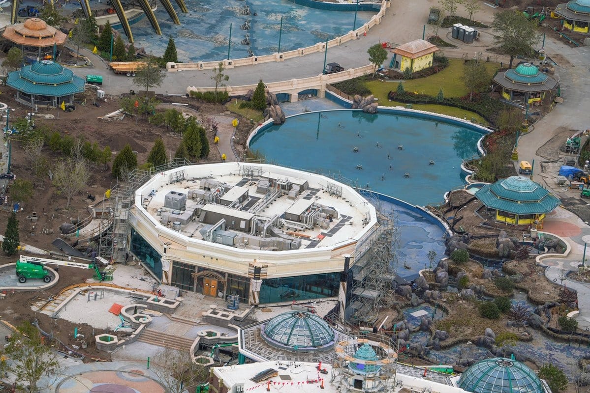 Aerial view of the Epic Universe theme park under construction, featuring a circular building with scaffolding, a large pond, pathways, and surrounding attractions.