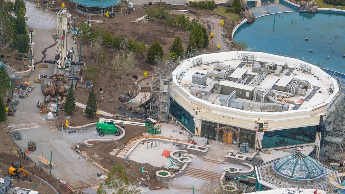Aerial view of an epic universe construction site with a circular building, machinery, and workers. Pathways and landscaped areas are visible.