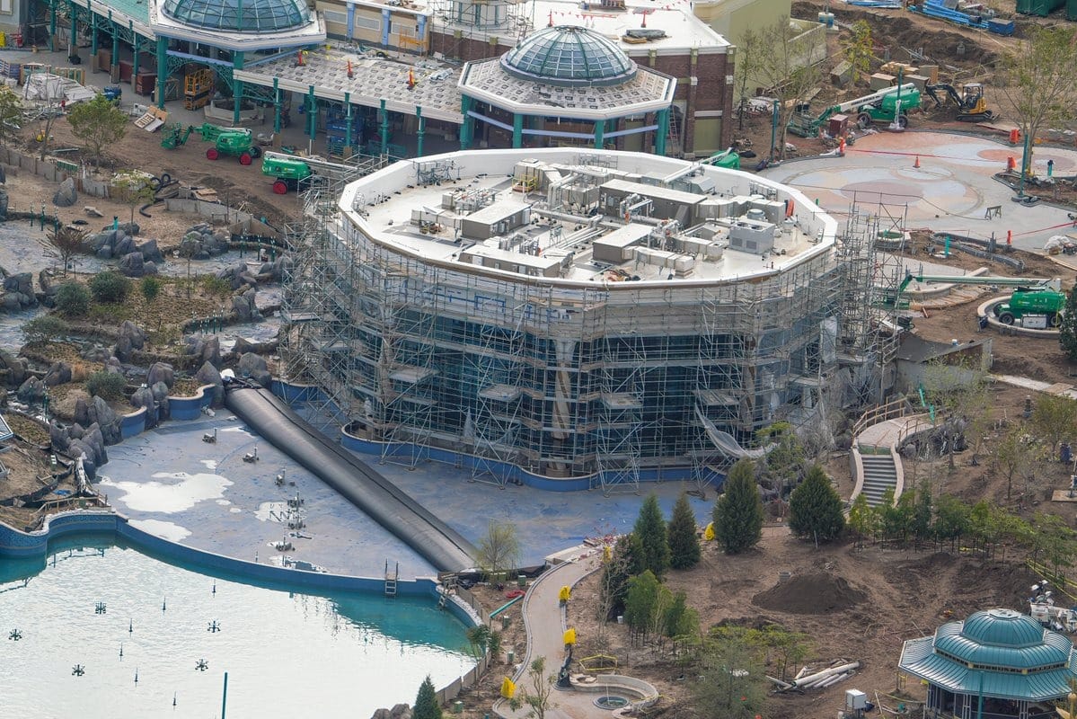 Aerial view of a construction site with a large building surrounded by scaffolding, evoking an epic universe in progress. Numerous construction vehicles and materials are visible. A pond with decorative elements graces the foreground.