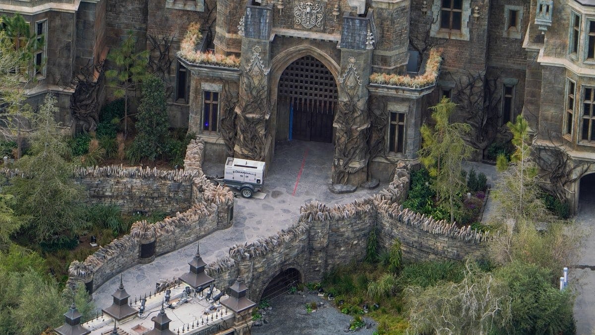 An aerial view reveals an epic universe where a stone castle with intricate designs sits surrounded by trees, while a vehicle is parked near its large arched entrance.