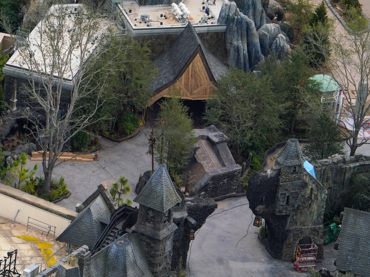 Aerial view of a medieval-themed park, part of the Epic Universe at Universal Orlando, featuring stone structures, a wooden-roofed building, pathways, and surrounding trees. Construction materials and equipment are visible nearby.