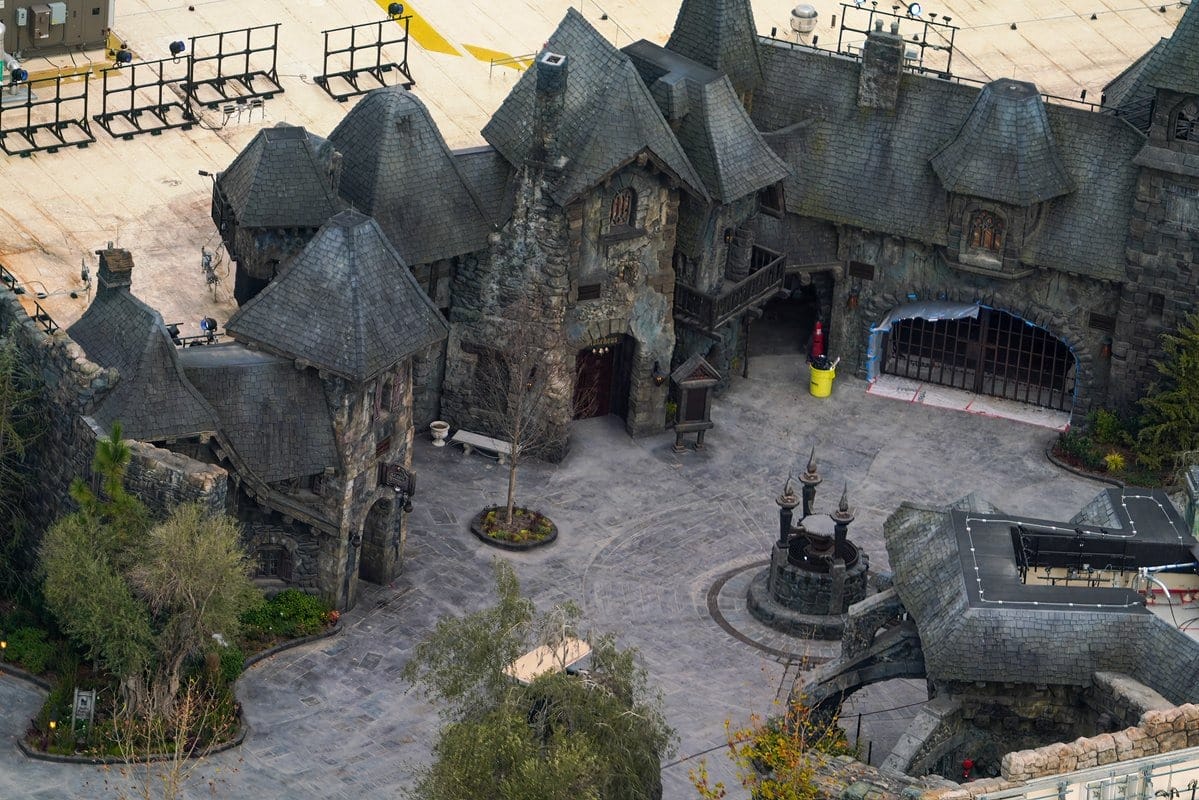 Aerial view of a castle-like structure in Epic Universe with stone walls and pointed roofs, featuring a courtyard with trees and a small fountain. Construction materials and equipment surround the theme park area.