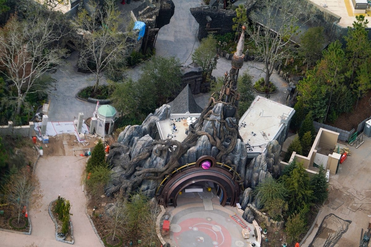 Aerial view of the Epic Universe attraction at Universal Orlando, featuring a building with twisted tree roots, a spire, and nearby trees. Pathways and construction areas are visible around the structure.