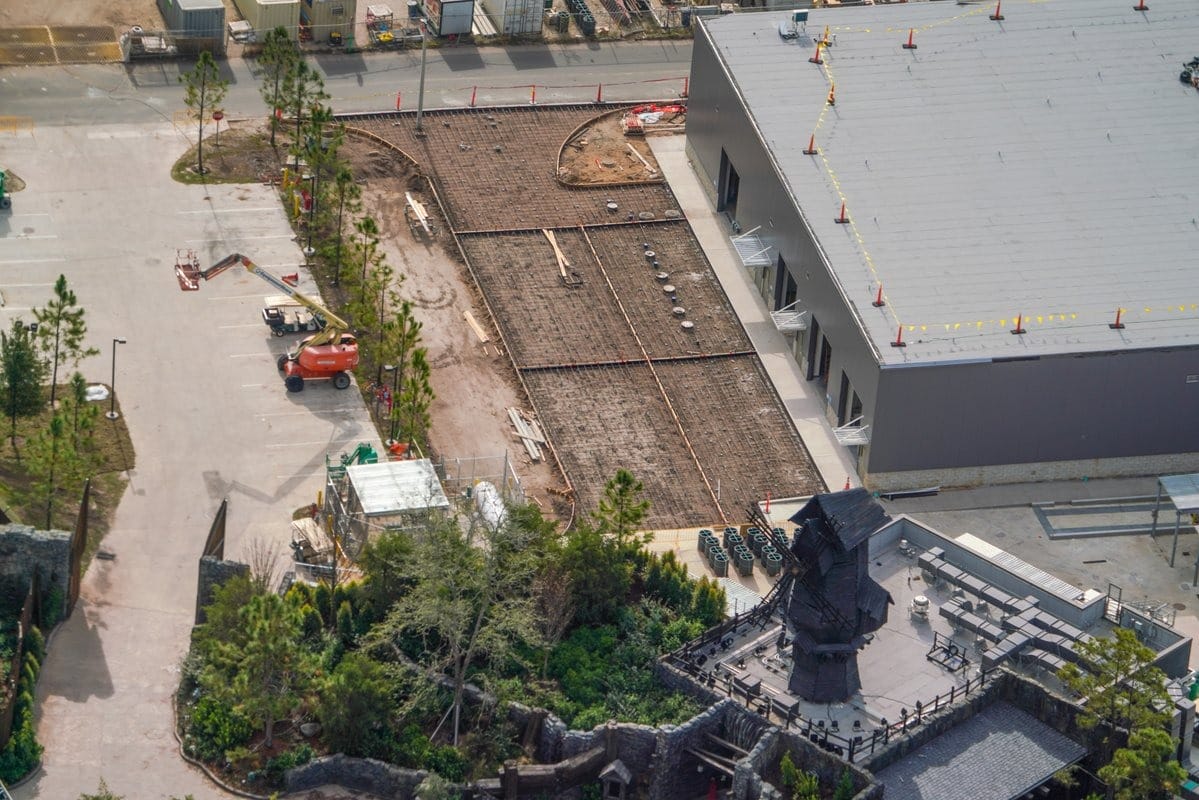 Aerial view of a construction site beside a large building, possibly part of an epic new addition to Universal's portfolio. The area is fenced, with construction materials and equipment visible. Nearby, there are trees and a small structure, adding intrigue to the evolving landscape.