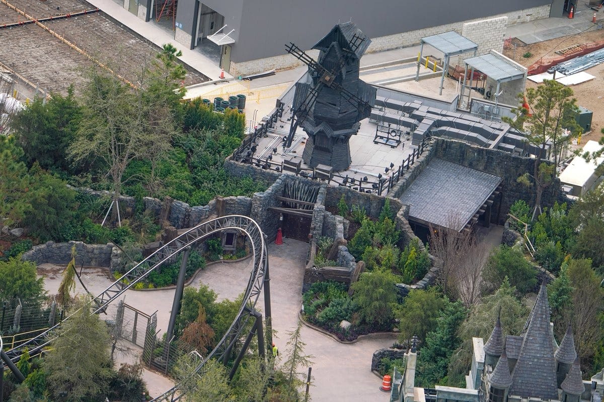 Aerial view of Epic Universe's theme park section featuring a thrilling roller coaster track, a dark windmill structure, and a winding path surrounded by lush trees and greenery.