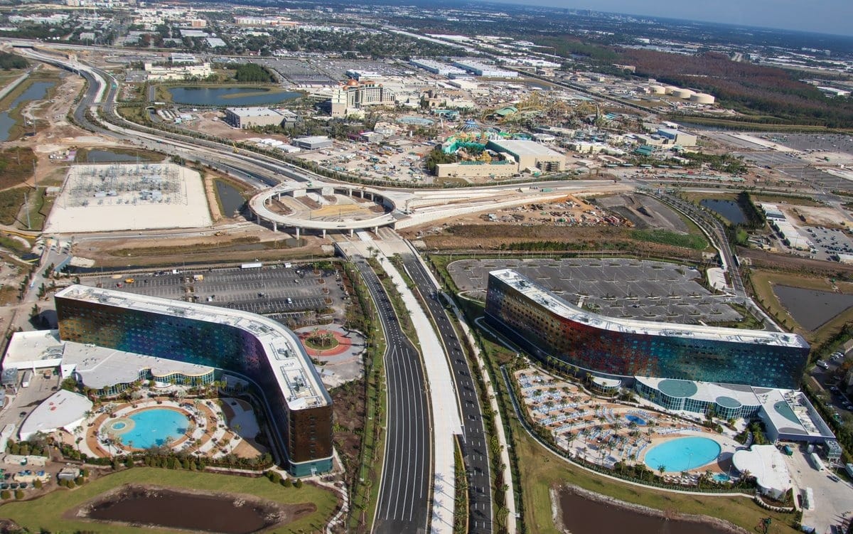 Aerial view of two large, curved buildings with colorful exteriors, surrounded by roads and parking lots. There are swimming pools and landscaped areas nearby.
