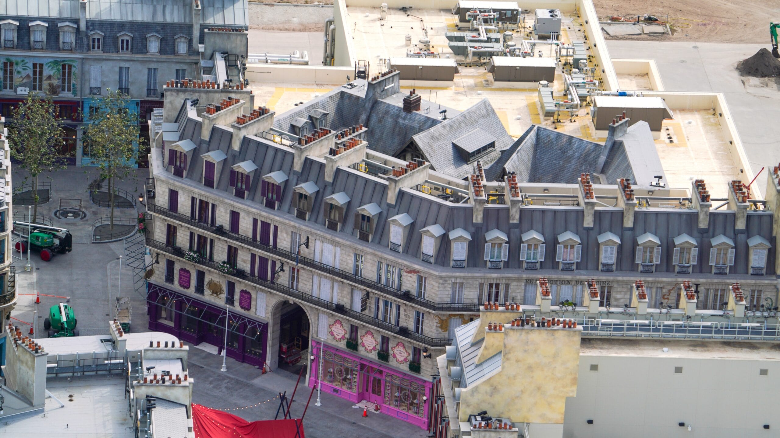 Aerial view of a building with a gray roof, many windows, and pink accents, surrounded by construction and machinery.