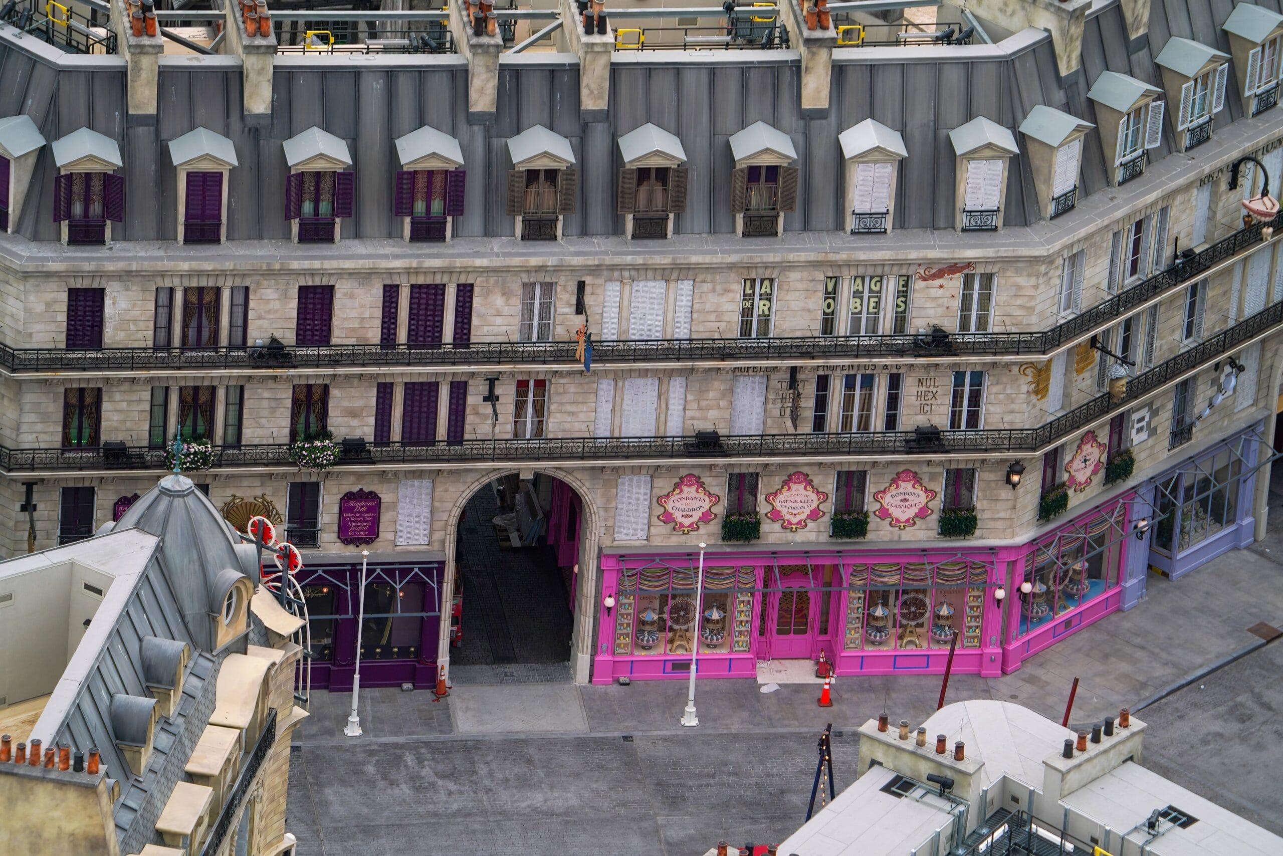 Aerial view of a street with a multi-story building featuring grey rooftops and purple and pink storefronts. There is a courtyard entrance in the center of the building.