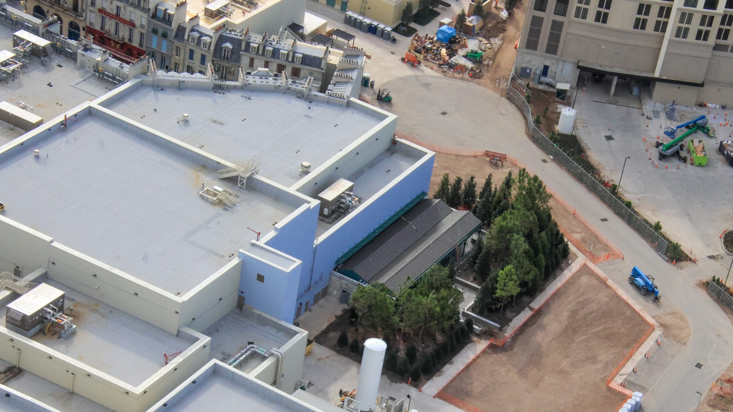Aerial view of a construction site with adjacent buildings, trees, and equipment. Workers and materials are visible throughout the area.