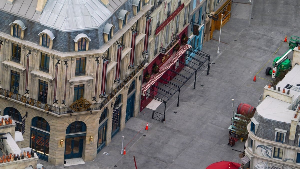 Aerial view of a city street with ornate buildings, an awning, and construction equipment, reminiscent of the grandeur surrounding Epic Universe attractions. Cones and barriers are set up along the sidewalk.