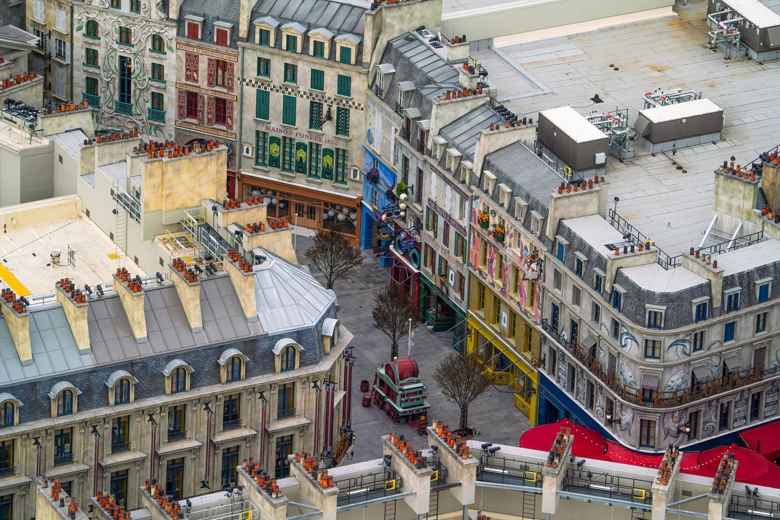 Aerial view of intricately detailed, colorful rooftops and building facades in a compact urban area, resembling the vibrant aesthetic of Epic Universe. It features a small plaza with decorative elements and a few bare trees, providing an immersive entertainment experience.