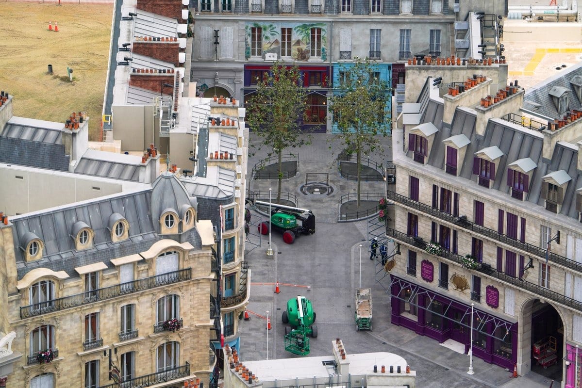 Bird's-eye view of urban buildings with a small courtyard. Two construction vehicles and several trees are in the courtyard. Surrounding buildings have distinct roofs and architectural details.