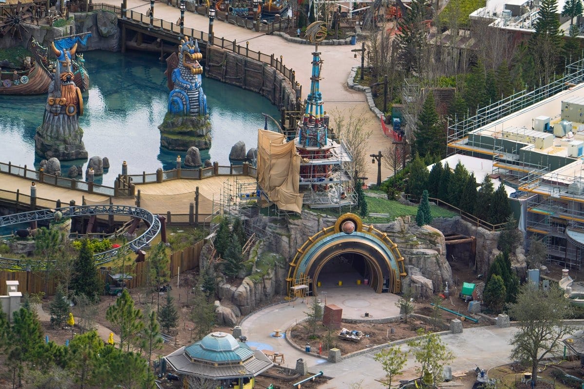 Aerial view of a themed amusement park area featuring construction zones, water with decorative statues, a roller coaster, and a partially built building with scaffolding.