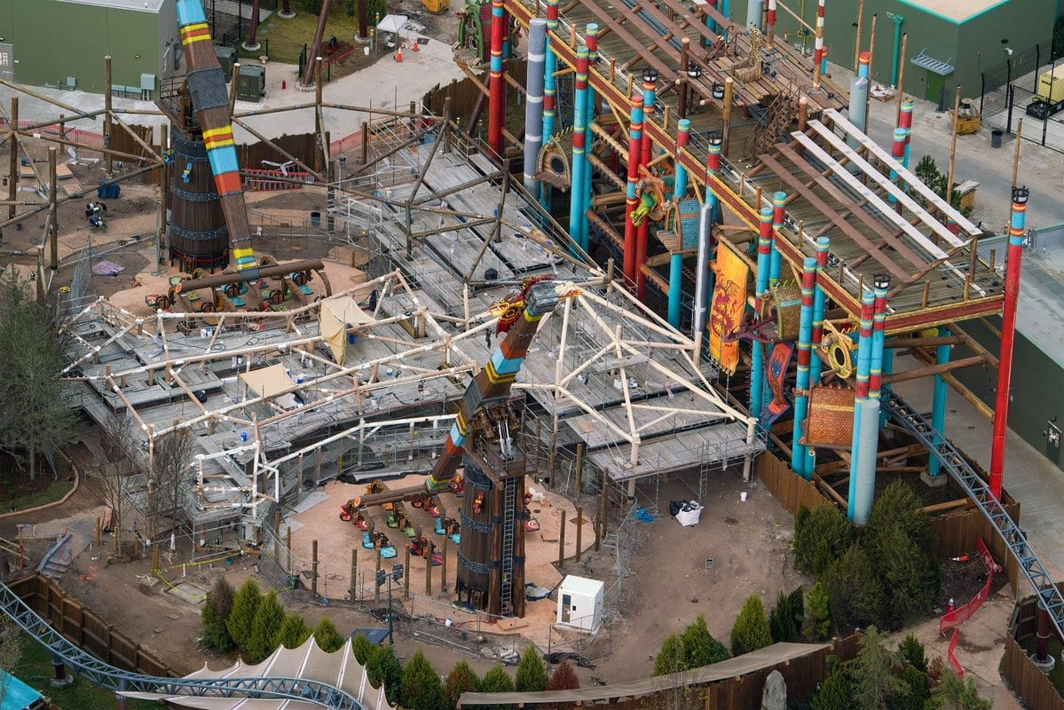 Aerial view of a theme park ride under construction, featuring vibrant colors, scaffolding, and decorative elements with towers and trees surrounding the area.