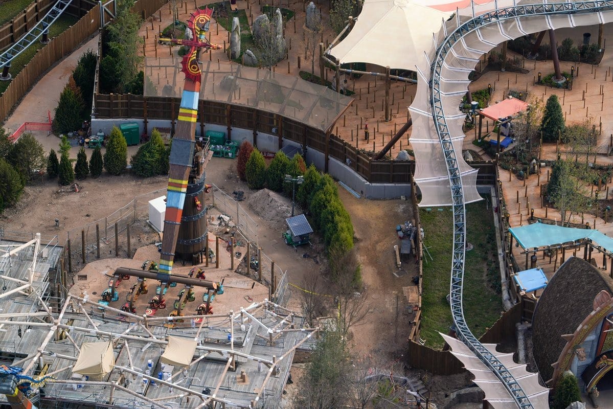 Aerial view of an amusement park ride with a spinning tower and nearby roller coaster tracks, surrounded by trees and pathways.