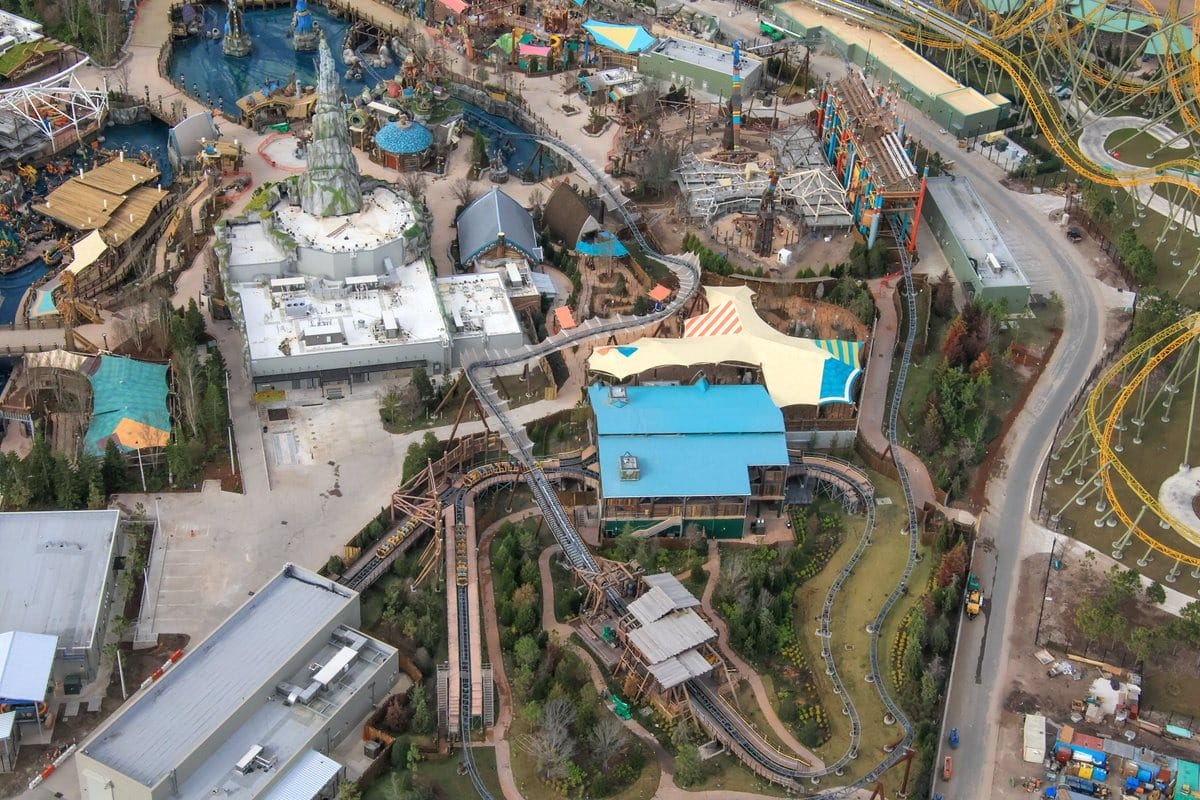 Aerial view of a theme park with roller coasters, pathways, buildings, and water features. Various colorful structures and landscaping are present throughout the park.