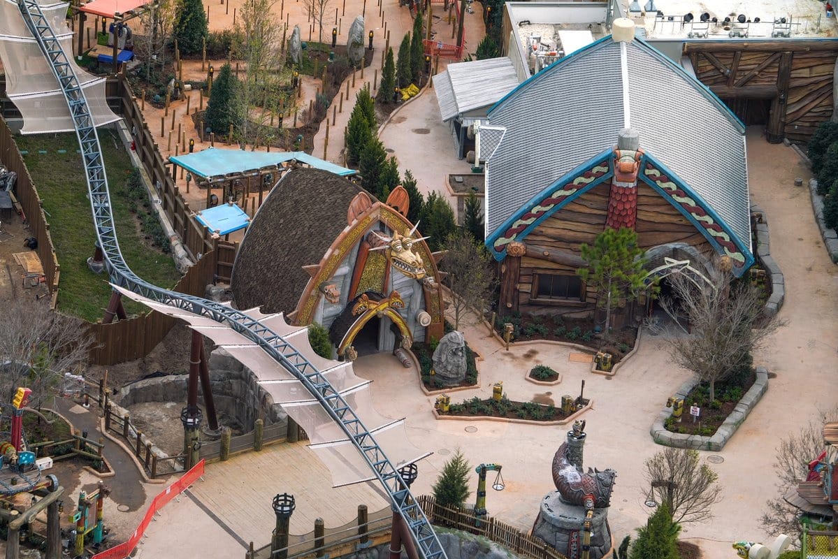 Aerial view of a theme park with a roller coaster track, a Viking-themed building, and a dragon head entrance. Paths and greenery surround the structures.