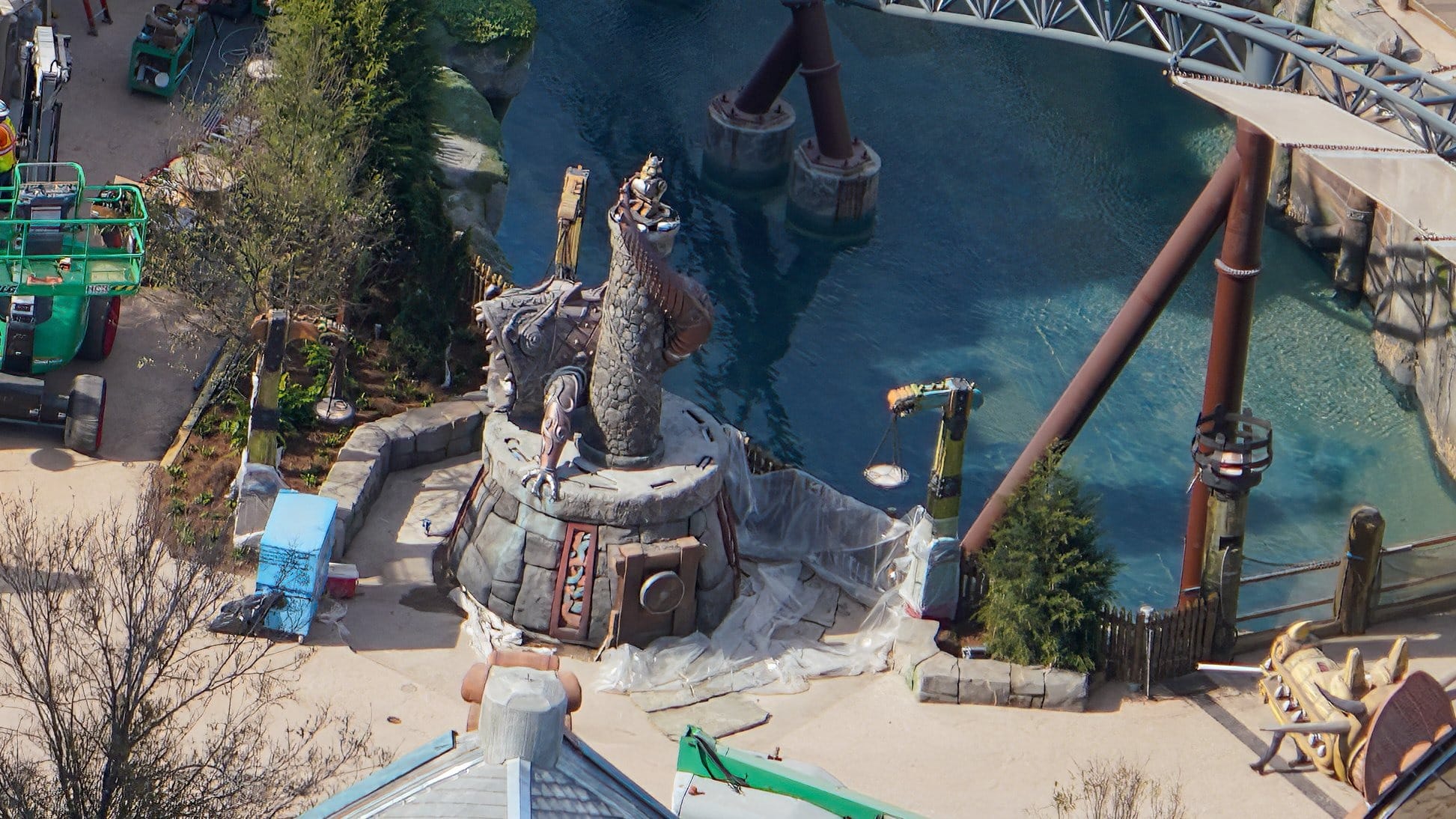 Aerial view of a large outdoor sculpture of a mythical creature by water, with construction equipment and greenery surrounding it.