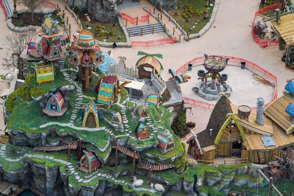 Aerial view of a colorful, intricate theme park with buildings resembling houses on a rocky landscape, paths, and a circular area with seating.