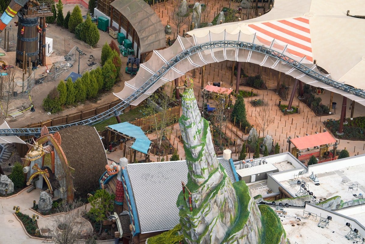 Aerial view of a theme park with a roller coaster track, rock formations, and themed structures. Trees and tents are visible in the background.