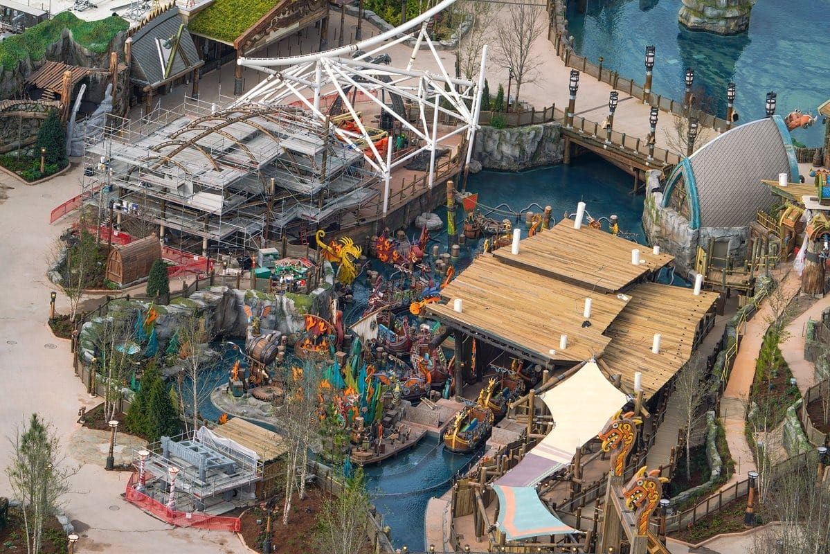 Aerial view of a themed amusement park featuring rides, colorful sculptures, waterways, and wooden structures. Scaffolding and construction materials are visible in certain areas.
