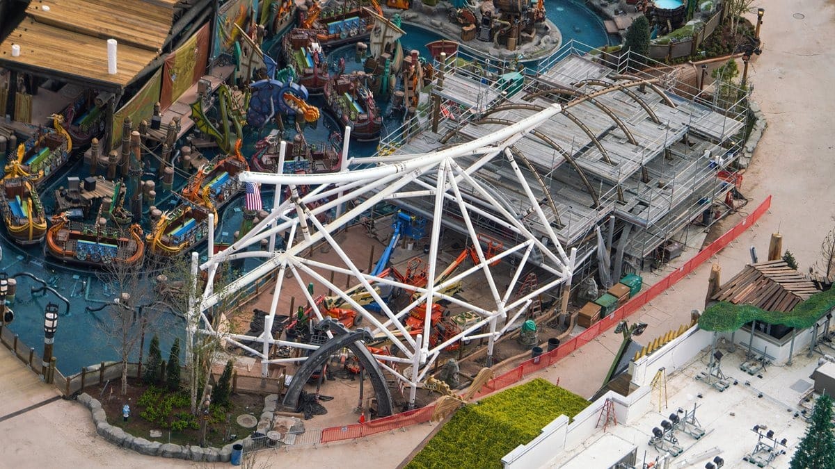 Aerial view of a roller coaster under construction at a theme park, surrounded by various themed props and pathways.