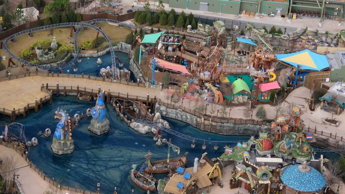 Aerial view of a theme park area with a water ride, colorful buildings, pathways, and decorative statues.