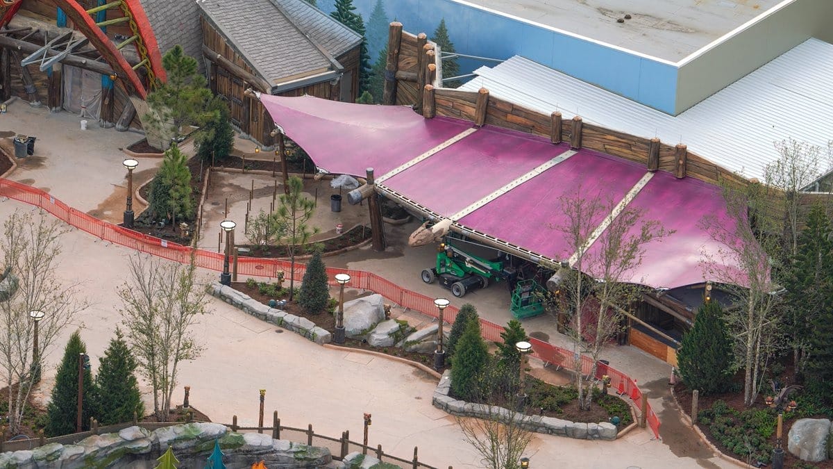 Aerial view of a theme park area with a large purple tent roof, surrounded by trees and pathways. Construction barriers are visible nearby.