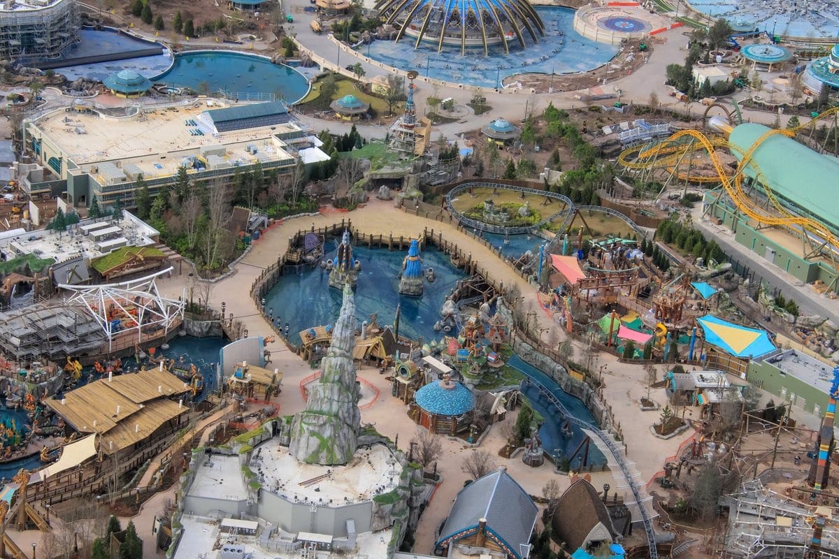 Aerial view of a theme park featuring various attractions, including water rides, a roller coaster, and themed buildings, surrounded by greenery and walkways.