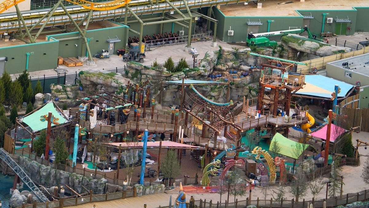 Aerial view of a colorful amusement park section with winding pathways, slides, climbing structures, and various play areas surrounded by green buildings and artificial rocks.