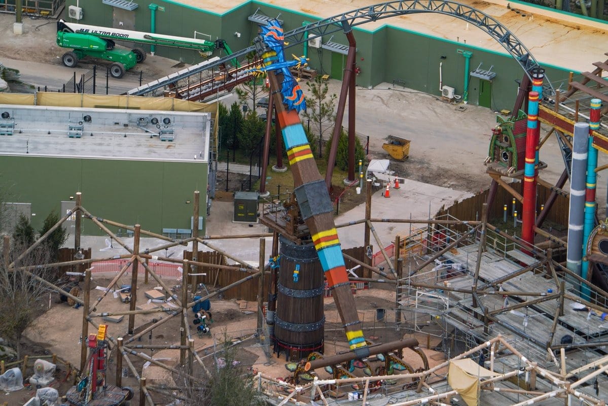 Aerial view of a theme park ride under construction, featuring a large swinging pendulum with vibrant blue, red, and yellow colors, surrounded by scaffolding and construction materials.