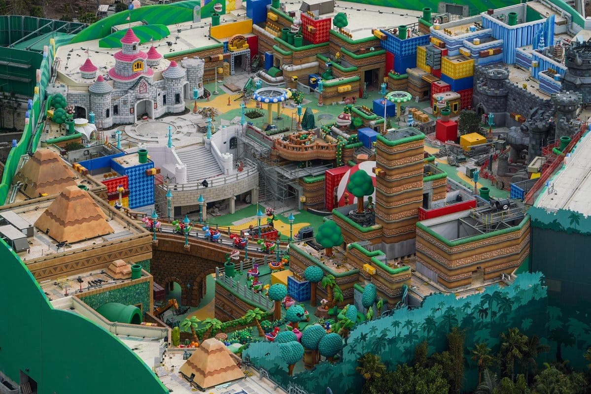 Aerial view of a colorful, multi-leveled theme park resembling a video game world, featuring castles, blocks, and various structures surrounded by greenery.