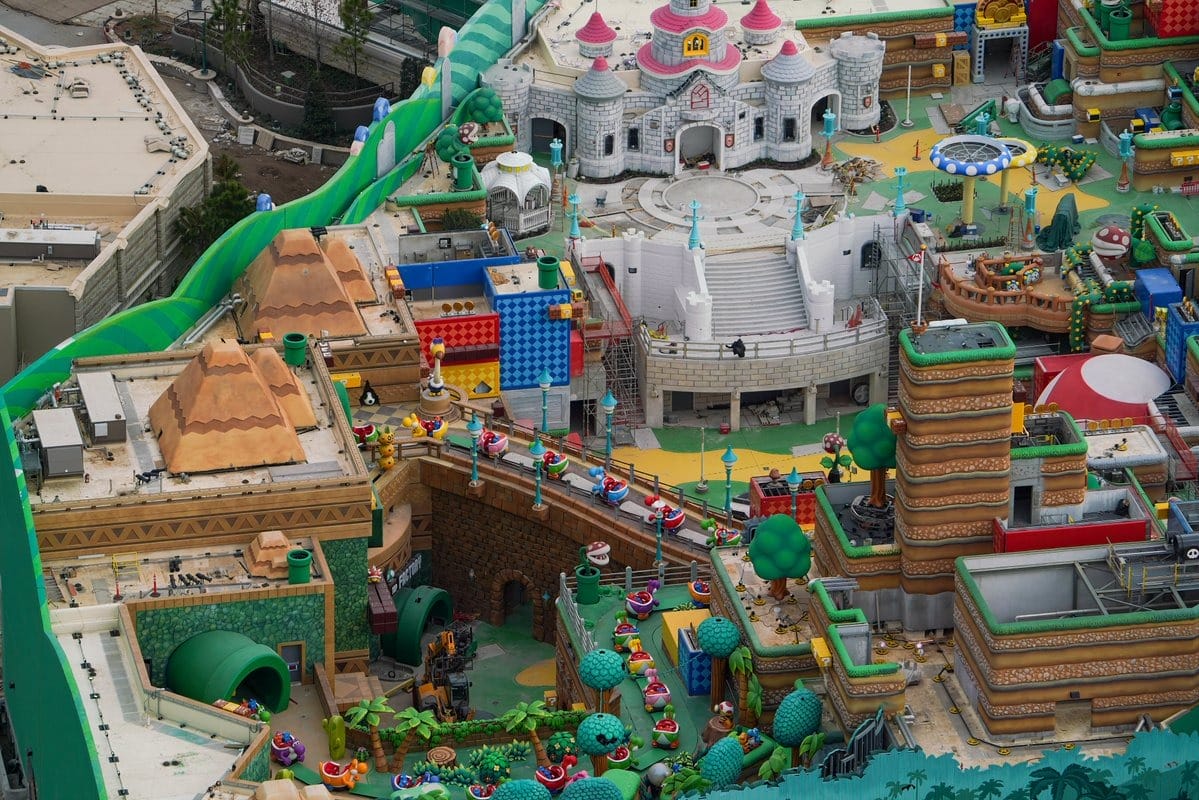 Aerial view of an amusement park section featuring a castle, colorful pathways, and themed structures with green and beige buildings surrounded by lush greenery and various themed elements.