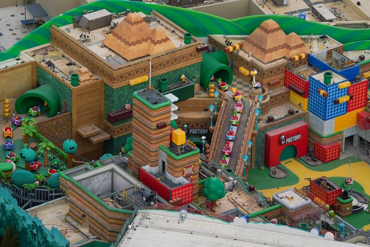 Aerial view of a colorful theme park area with block-like structures, characters, and a factory entrance, resembling a video game scene with pyramids and green pipes.