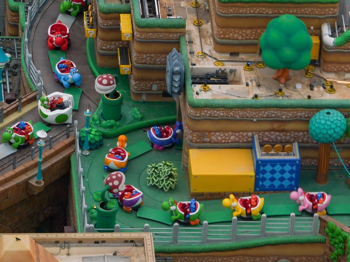 Aerial view of a colorful theme park ride featuring small cars shaped like red turtles winding through a landscaped area with green structures and various themed elements.