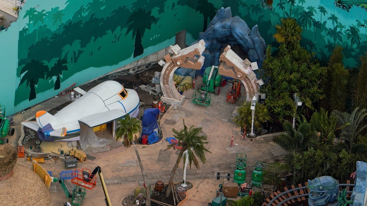 Aerial view of a theme park under construction with a white airplane, stone arches, and lush greenery. Equipment and construction materials are visible around the site.