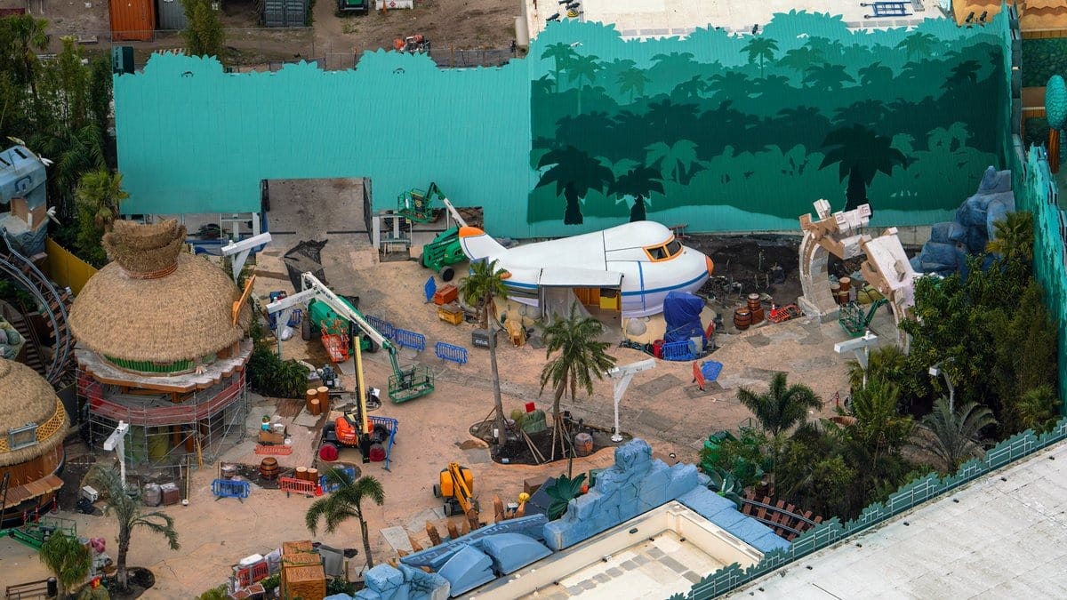 Aerial view of a theme park under construction with a stylized propeller plane, palm trees, and circular straw-roofed buildings surrounded by construction equipment and cranes.