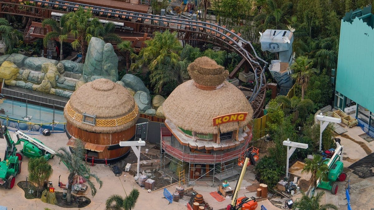 Aerial view of a theme park under construction featuring two straw-roofed huts labeled "KONG," surrounded by construction vehicles, a winding track, and various landscaping elements.