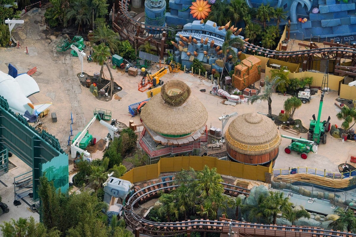 Aerial view of a theme park construction site with tropical-themed structures, foliage, and various construction vehicles.