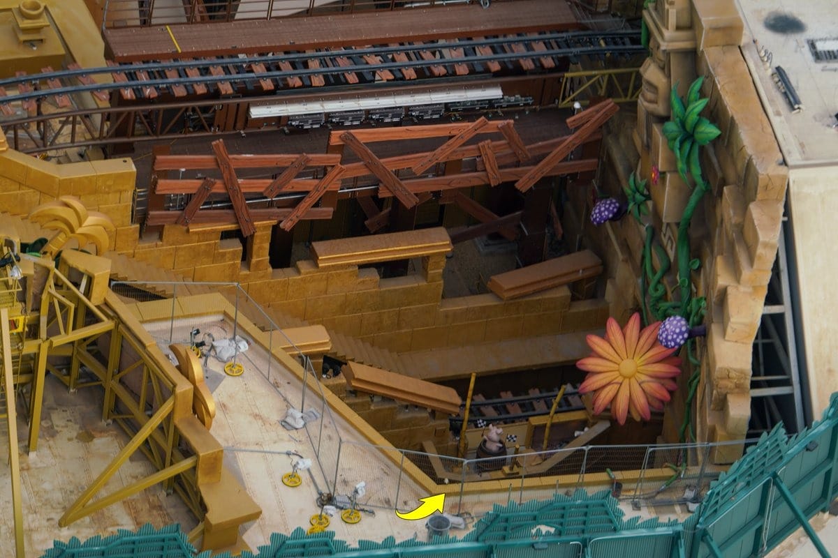 Construction site with wooden structures, a large decorative flower, and green fencing. Partially built track and pathways are visible.