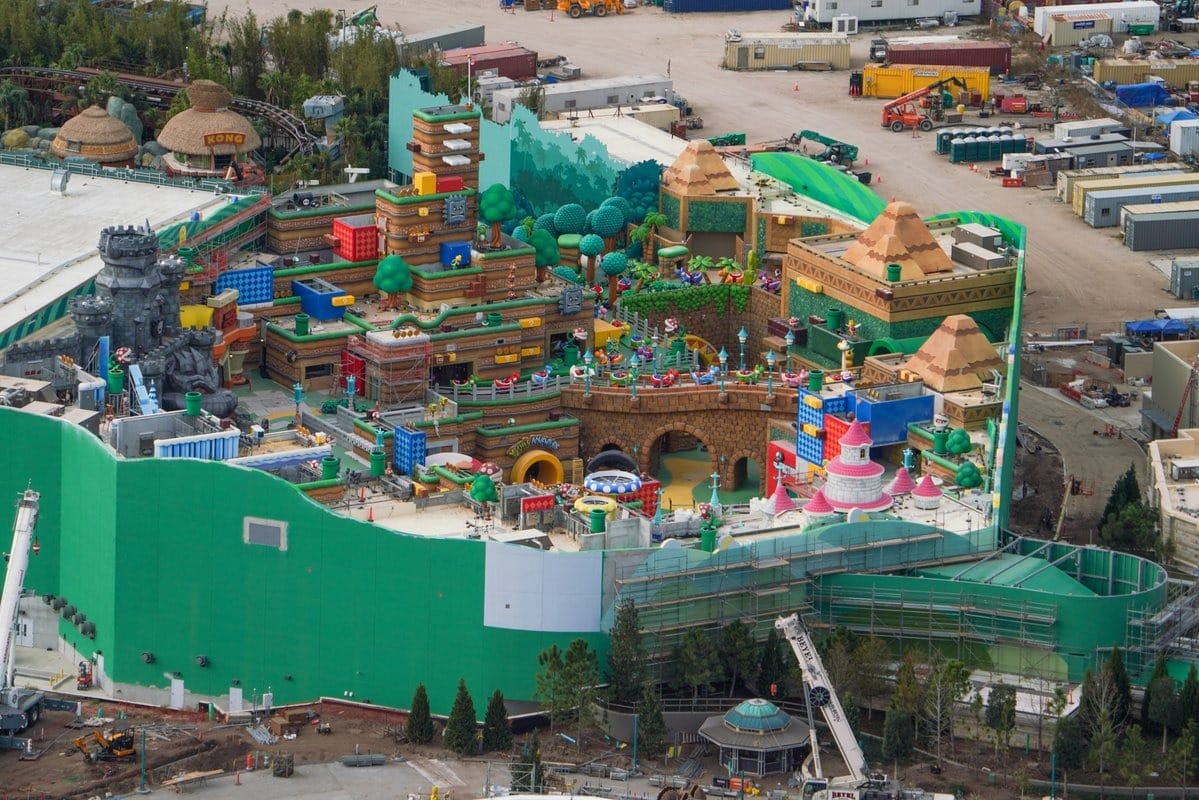 Aerial view of a colorful amusement park under construction, featuring structures resembling video game elements, surrounded by cranes and construction vehicles.