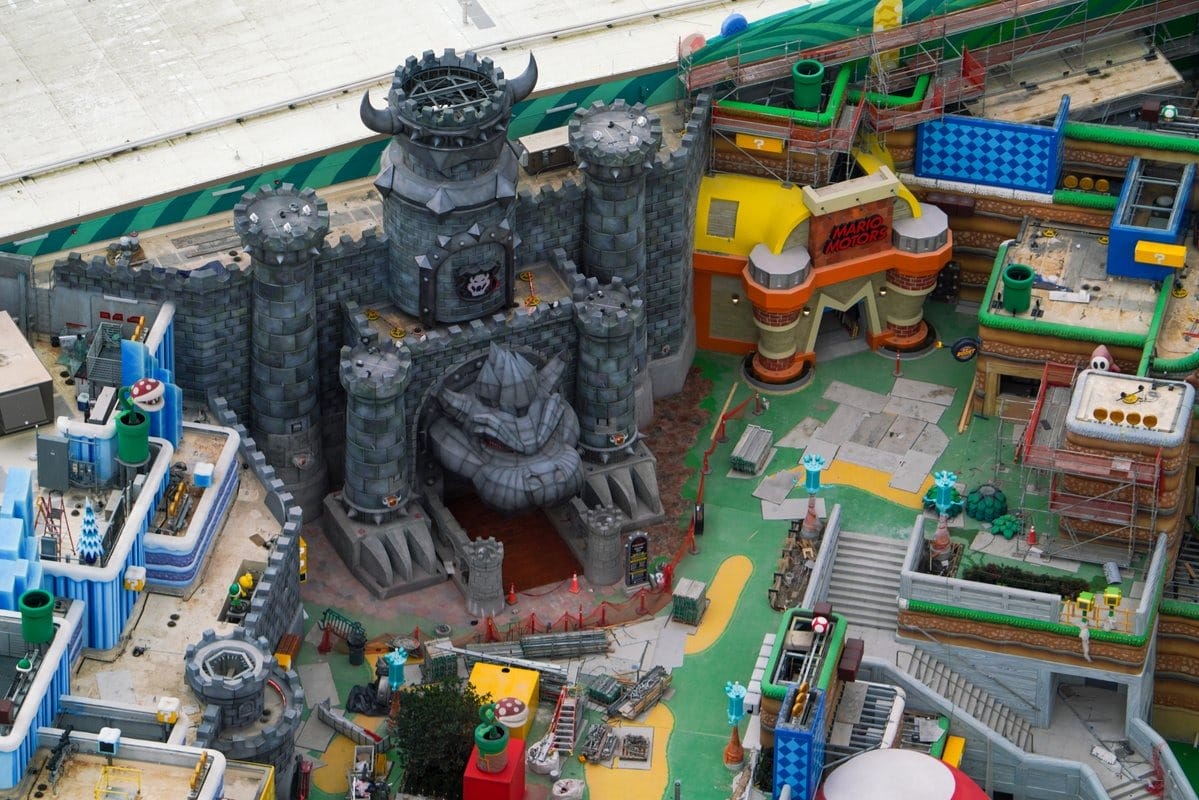 Aerial view of a theme park with a gray castle featuring a large face, surrounded by colorful structures and walkways. Construction materials are visible.