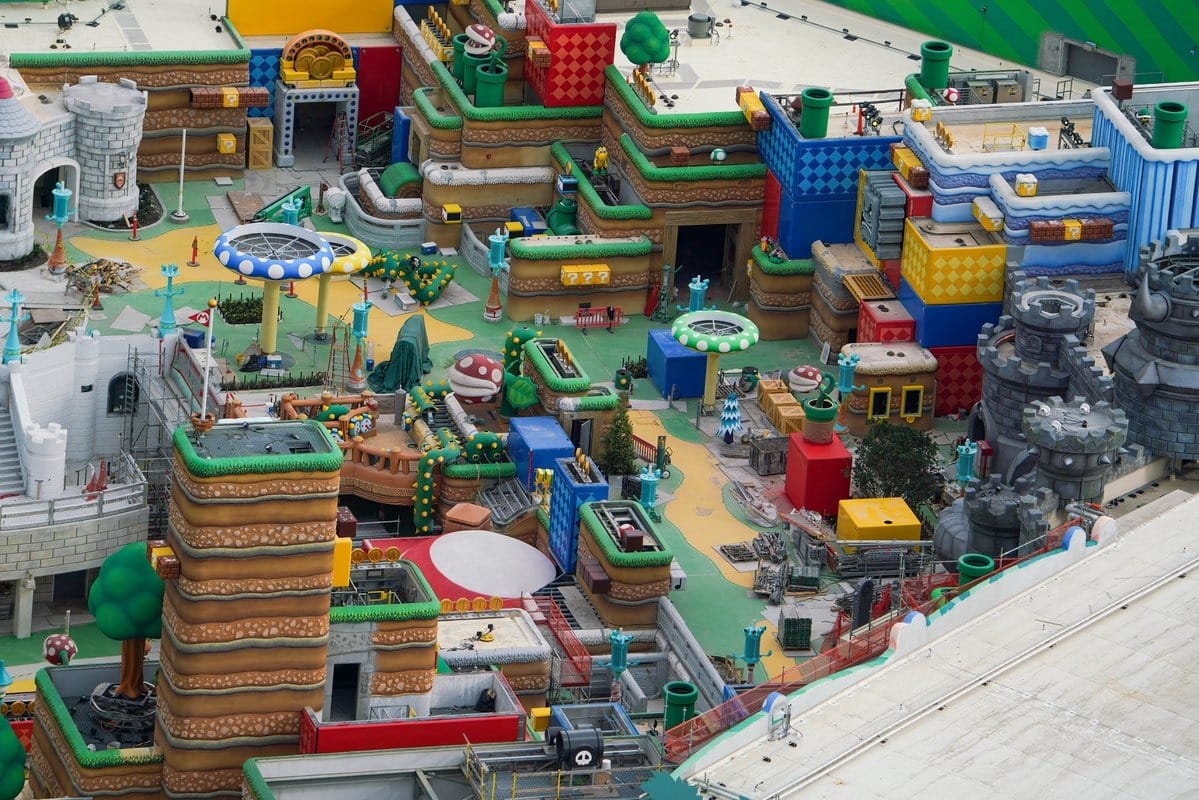 Aerial view of a colorful, multi-level theme park area with various structures resembling a video game landscape, featuring mushrooms, castles, and green pipes.