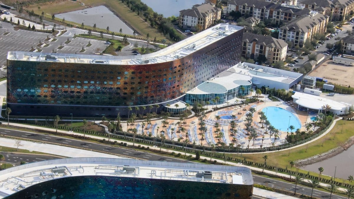 Aerial view of a modern, curved hotel building with circular windows, adjacent to a large pool and landscaped area, surrounded by roads, parking lots, and residential complexes.