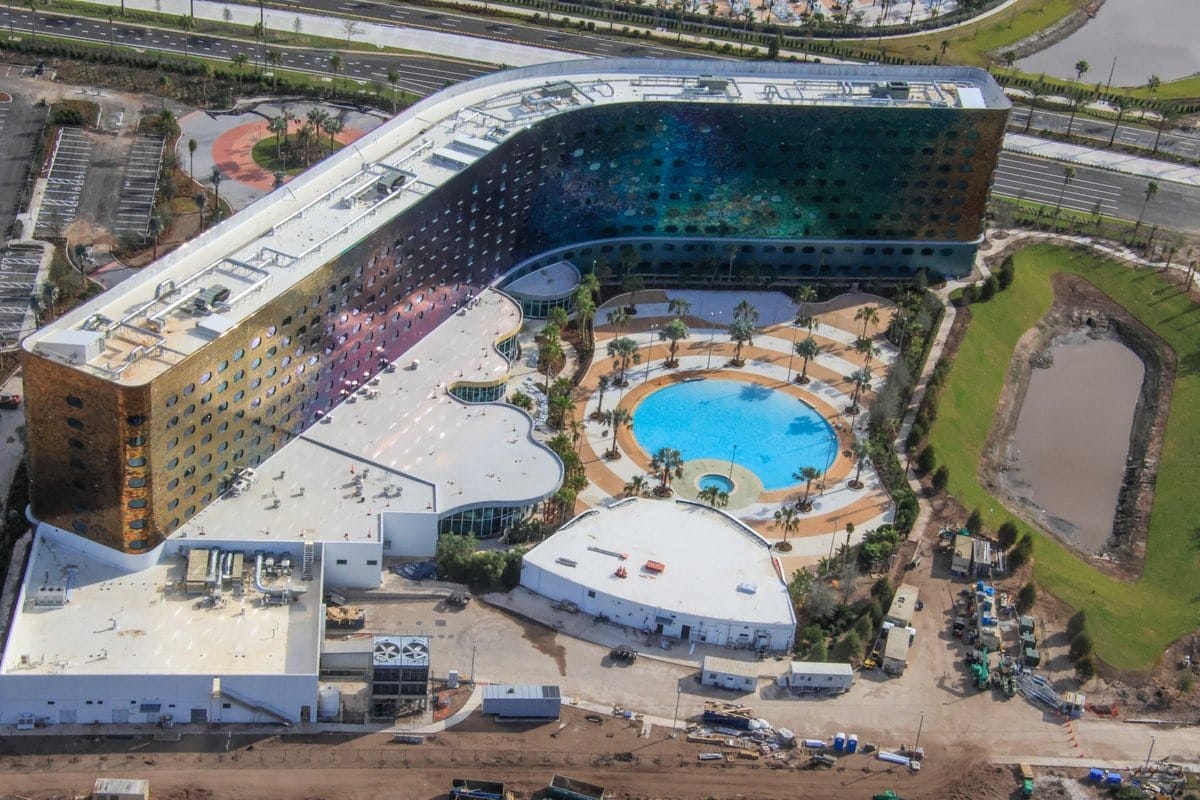 Aerial view of a large, modern hotel complex with a curved design, featuring a central courtyard with a circular pool and palm trees. Surrounding areas show construction and landscaping in progress.