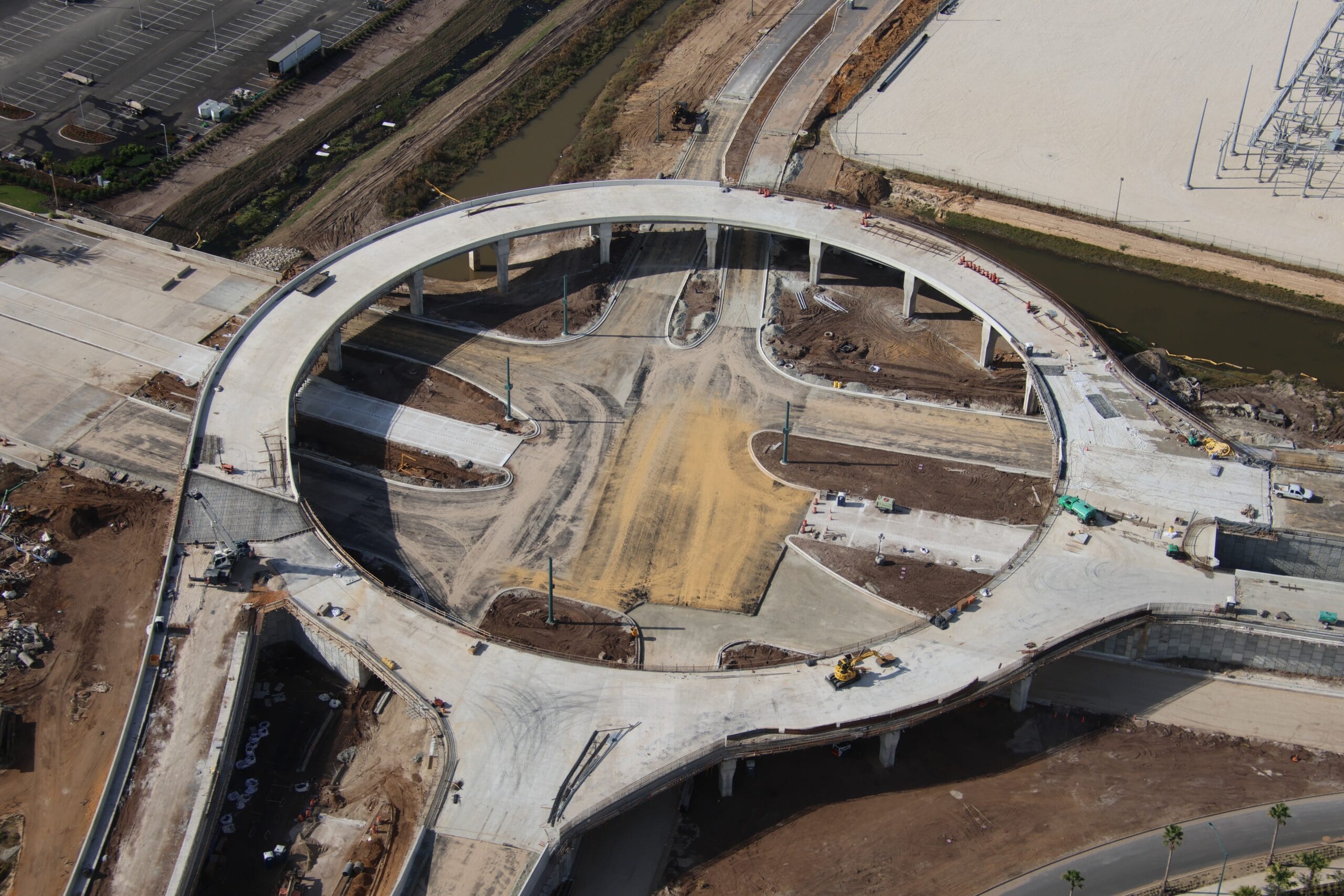 Aerial view of an unfinished circular highway overpass under construction, resembling the grand designs of an Epic Universe theme park, surrounded by various construction materials and a nearby road.