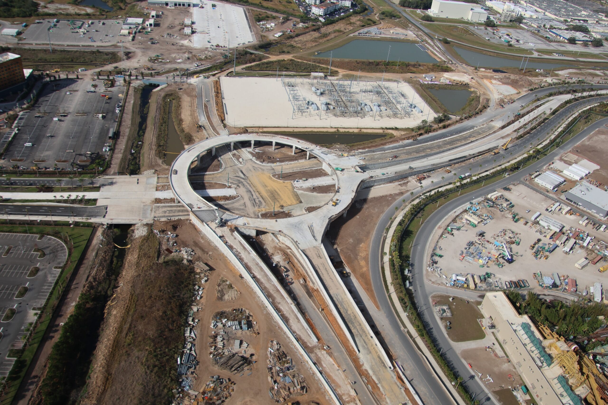 An aerial view of a highway interchange under construction near Orlando reveals a circular road with multiple entry and exit ramps, surrounded by construction materials. Nearby buildings hint at the future development of the Epic Universe theme park.