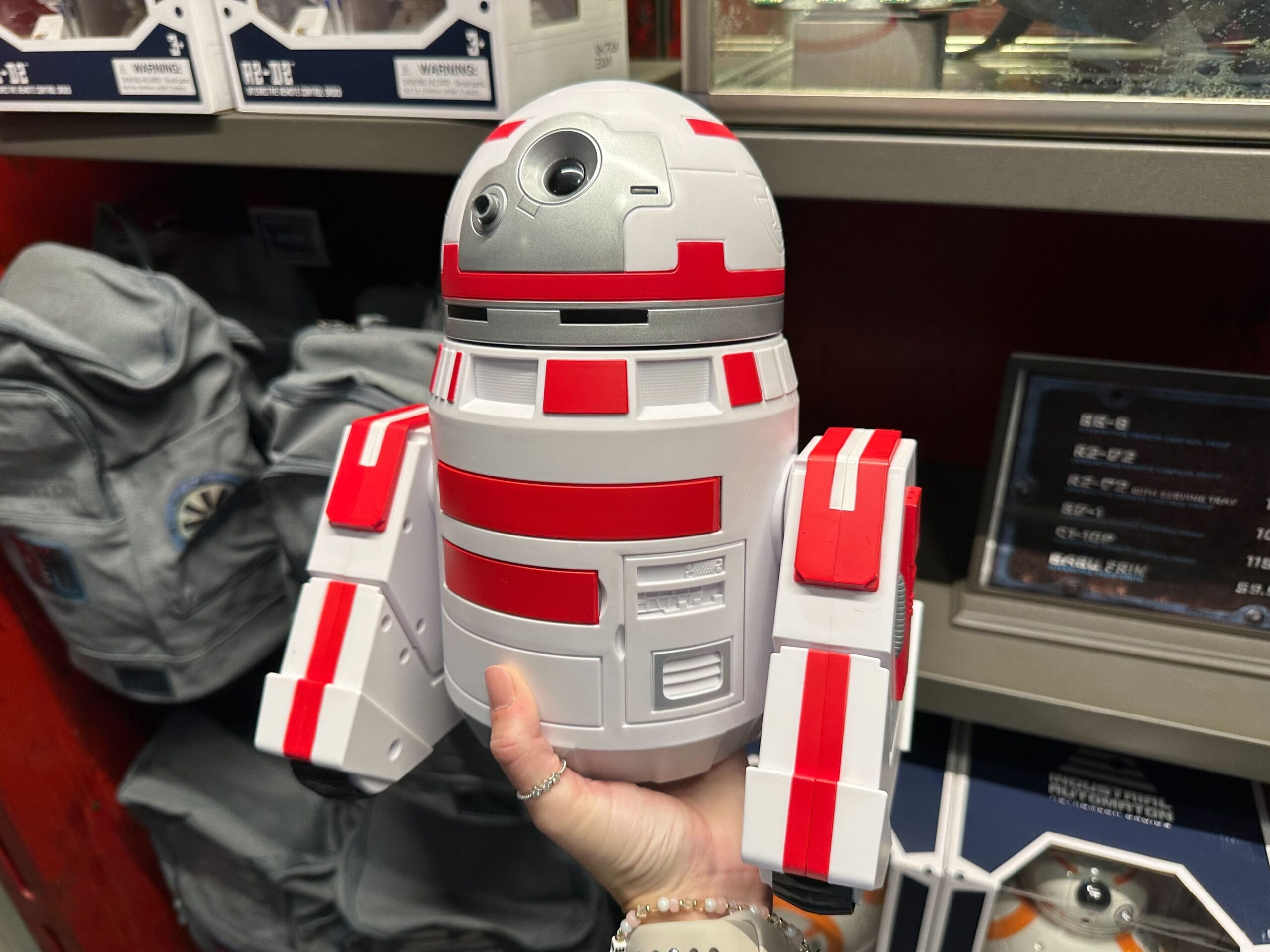A person holding a red-and-white robot toy resembling a popular sci-fi droid, displayed in a store with backpacks and other items in the background.