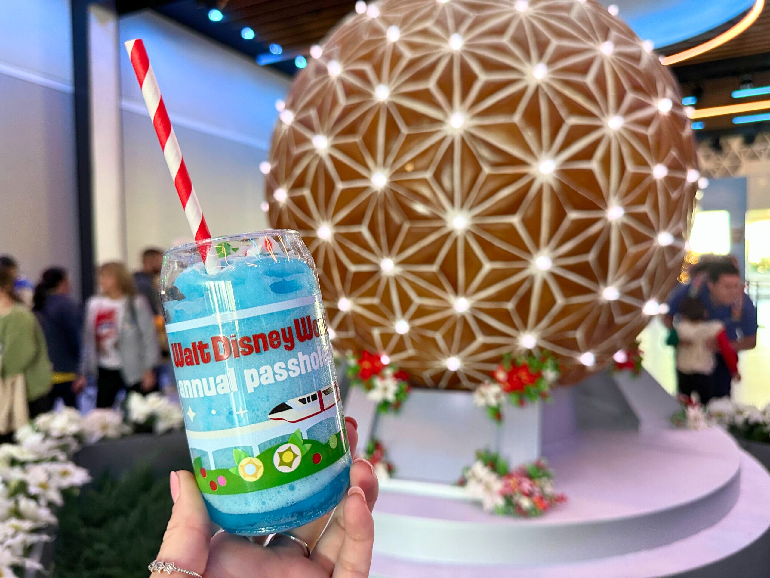 A person holds a themed cup with "Walt Disney World Annual Passholder" text. In the background, a decorative sphere is lit up with white lights.