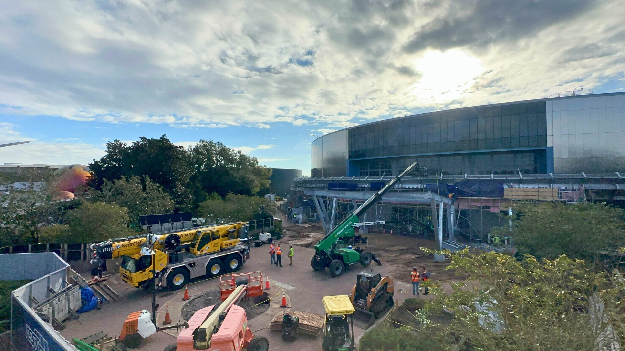 Construction site with cranes and vehicles working outside a large building under cloudy skies, surrounded by trees and orange safety cones. Several workers are visible on the ground.